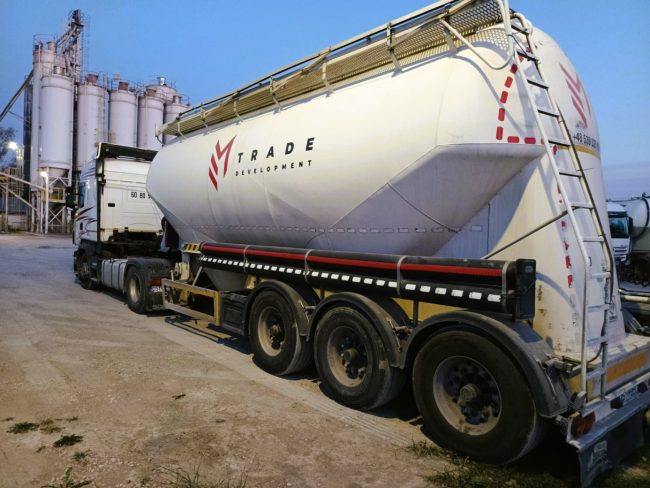 Ciężarówka z naczepą typu silos firmy EM Trade Development, stojąca na terenie bazy logistycznej obok silosów magazynowych, przeznaczona do transportu materiałów sypkich, takich jak nowoczesne hydrauliczne spoiwa drogowe.