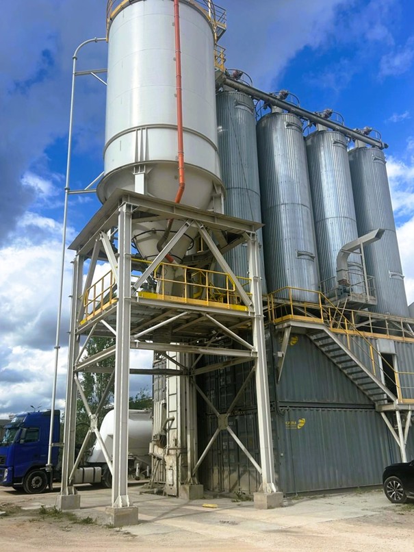 Wysoki, biały silos magazynowy wsparty na stalowej konstrukcji oraz rząd mniejszych, metalowych zbiorników na terenie zakładu produkcyjnego, służące do przechowywania i dozowania materiałów sypkich, takich jak hydrauliczne spoiwa drogowe.